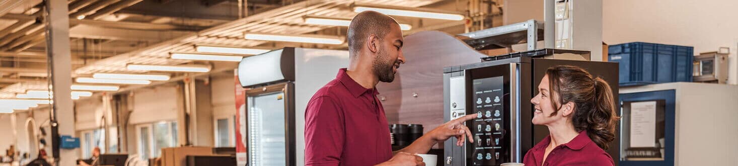 Zwei Mitarbeiter trinken Kaffee neben Kaffeevollautomat in einer Werkstatt.