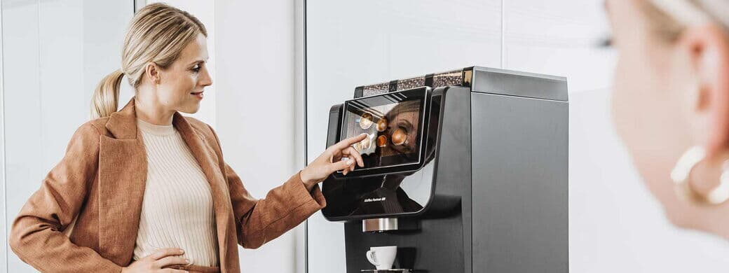 Eine Frau in eleganter Bürokleidung bedient einen Kaffeevollautomaten von Kaffee Partner im Büro. Eine weitere Frau ist im Vordergrund leicht unscharf zu sehen.