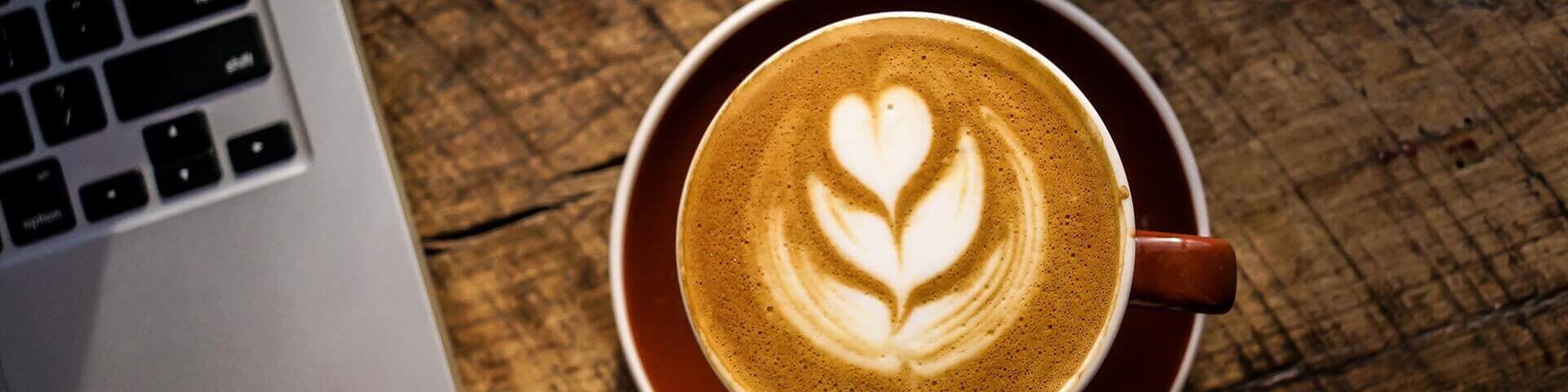 Eine Tasse Kaffee mit einer Blume aus Milchschaum steht neben einem Laptop auf einem Holztisch