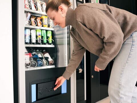 Person entnimmt Produkt aus Ausgabefach eines Snackautomaten