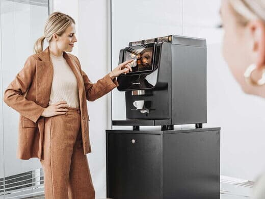 Eine Frau in eleganter Bürokleidung bedient einen Kaffeevollautomaten von Kaffee Partner im Büro. Eine weitere Frau ist im Vordergrund leicht unscharf zu sehen.